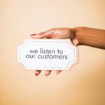 A hand holding a sign saying 'we listen to our customers' on a neutral background.